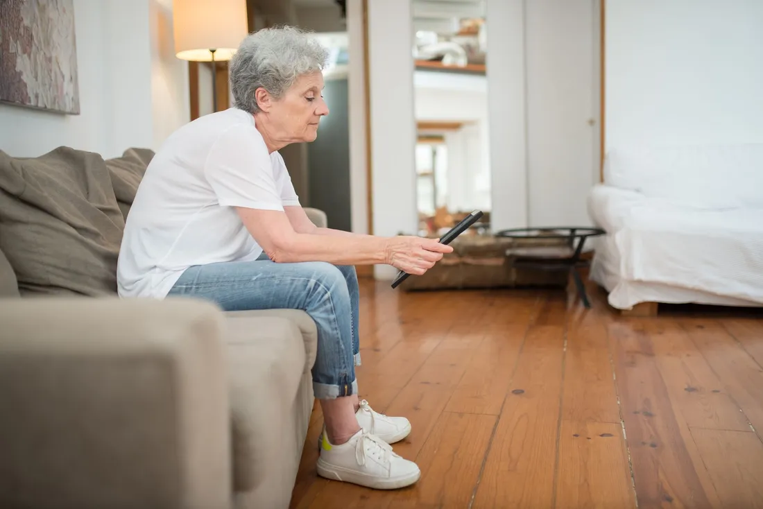 Femme âgée assise sur son canapé, dans un salon au parquet en bois