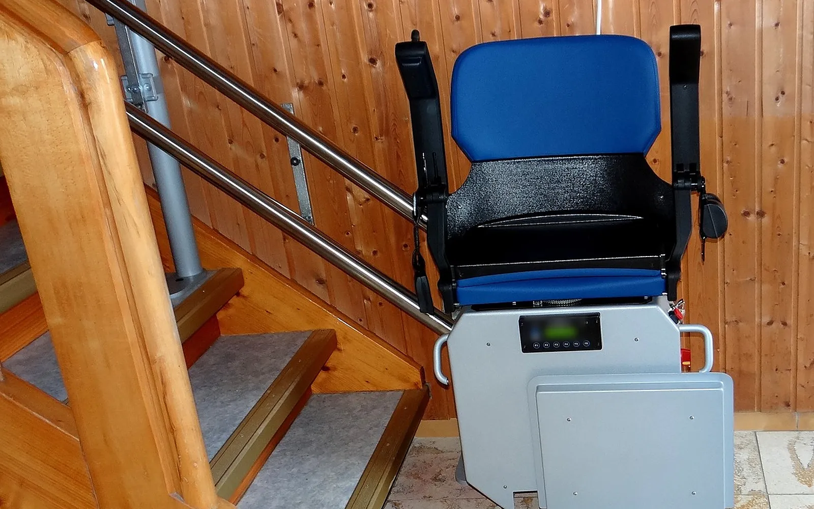 Monte-escalier électrique replié au pied d'un escalier droit en bois : siège bleu, accoudoirs et repose-pieds relevés, rail double en inox le long des marches